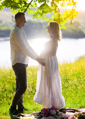 Beautiful young pregnant woman with her husband in the forest at sunset on a picnic sitting on the grass