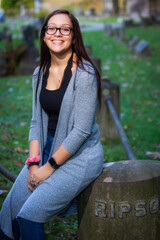 portrait of a pretty teenaged girl with dark hair and glasses seated in a cemetery