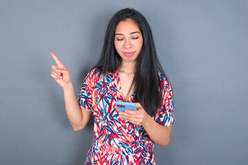 Smiling Young latin woman wearing colorful dress over grey backgrogund pointing finger at blank space holding phone in one hand