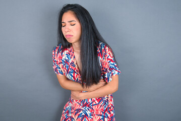 Young latin woman wearing colorful dress over grey backgrogund got stomachache