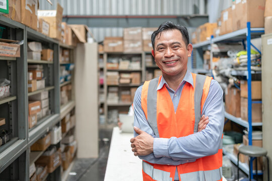 Smiling Asian senior male supervisor in safety vest standing arm crossed at warehouse office