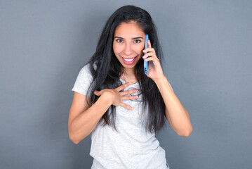 Smiling young beautiful brunette woman wearing white t-shirt over grey background  talks via cellphone, enjoys pleasant great conversation. People, technology, communication concept