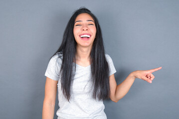 young beautiful brunette woman wearing white t-shirt over grey background laughs happily points away on blank space demonstrates shopping discount offer, excited by good news or unexpected sale.