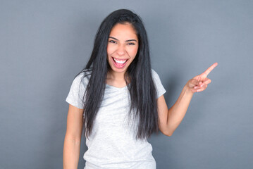 young beautiful brunette woman wearing white t-shirt over grey background points aside on copy blank space. People promotion and advertising concept