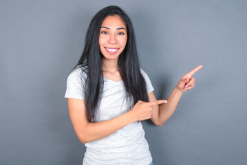 young beautiful brunette woman wearing white t-shirt over grey background points at copy space indicates for advertising gives right direction