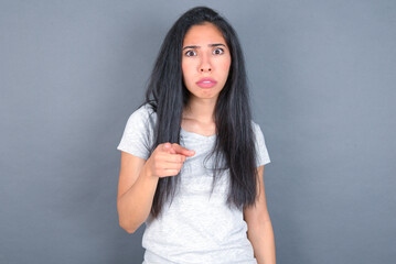 Shocked young beautiful brunette woman wearing white t-shirt over grey background points at you with stunned expression