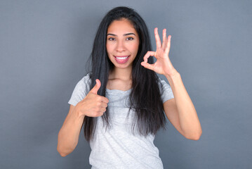 young beautiful brunette woman wearing white t-shirt over grey background smiling and looking happy, carefree and positive, gesturing victory or peace with one hand