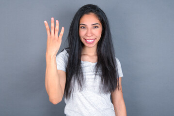 young beautiful brunette woman wearing white t-shirt over grey background smiling and looking friendly, showing number four or fourth with hand forward, counting down