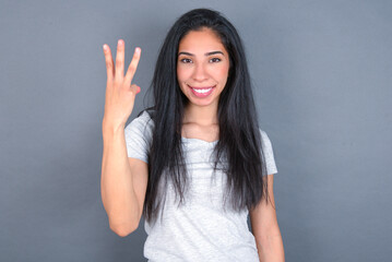 young beautiful brunette woman wearing white t-shirt over grey background smiling and looking friendly, showing number three or third with hand forward, counting down