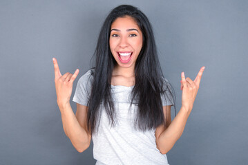 young beautiful brunette woman wearing white t-shirt over grey background makes rock n roll sign looks self confident and cheerful enjoys cool music at party. Body language concept.