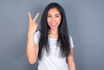 young beautiful brunette woman wearing white t-shirt over grey background smiling and looking friendly, showing number two or second with hand forward, counting down