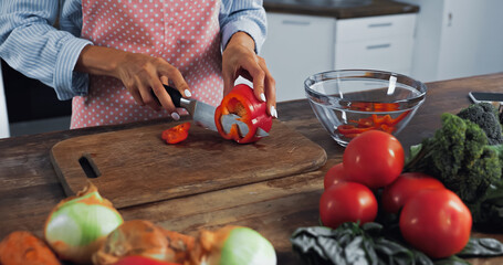 cropped view of woman cutting bell pepper near bowl and different vegetables on worktop.