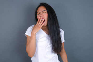 Sleepy young beautiful brunette woman wearing white t-shirt over grey background yawning with messy hair, feeling tired after sleepless night, yawning, covering mouth with palm.