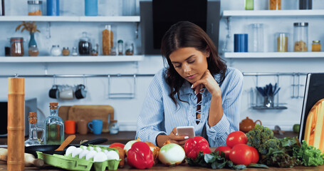 woman using smartphone near various ingredients and cookbook on kitchen table.