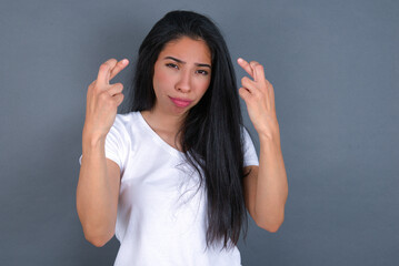 young beautiful brunette woman wearing white t-shirt over grey background holding fingers crossed...