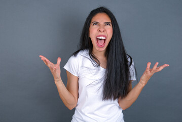 young beautiful brunette woman wearing white t-shirt over grey background crying and screaming....