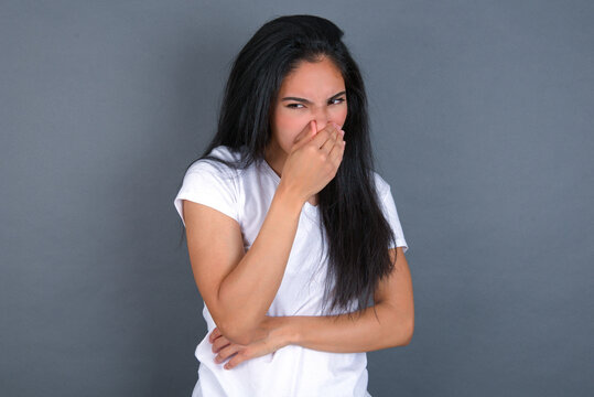 young beautiful brunette woman wearing white t-shirt over grey background, holding his nose because of a bad smell.