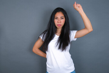 young beautiful brunette woman wearing white t-shirt over grey background feeling serious, strong and rebellious, raising fist up, protesting or fighting for revolution.