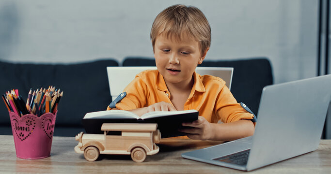 boy looking at notebook near laptop and wooden toy car on desk.