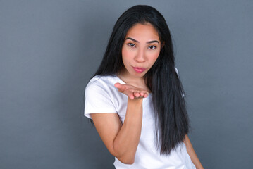 Obraz premium young beautiful brunette woman wearing white t-shirt over grey background looking at the camera blowing a kiss with hand on air being lovely. Love expression.