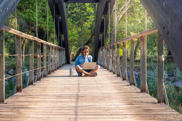 Young man working every day in a different relaxed place sunbathing sitting on a wooden bridge in...