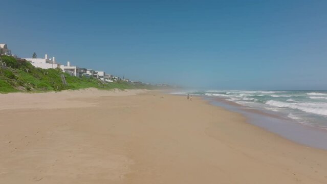 Couple strolling on beach along rolling waves. Forwards fly along coast, houses near sea. South Africa