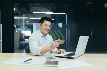 Fototapeta na wymiar Happy Asian businessman working in modern office, man making online money transfer, using laptop and holding bank credit card