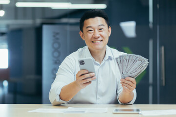 Naklejka na ściany i meble Successful asian businessman working in office, man smiling and looking at camera, holding phone and money dollars cash