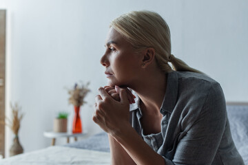 side view of worried blonde woman with menopause looking away in bedroom. - obrazy, fototapety, plakaty
