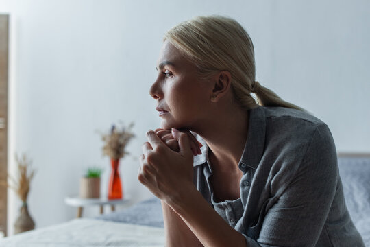 side view of worried blonde woman with menopause looking away in bedroom.