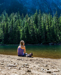 An adventurous athletic female hiker sitting on the shore of an alpine lake in the Pacific Northwest with her French Bulldog.