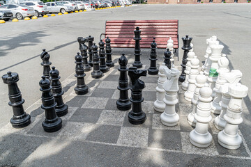 A chessboard on asphalt with white and black chess pieces for playing and competing in a chess battle on the square in front of the City Administration building near the park and a bench for fans.