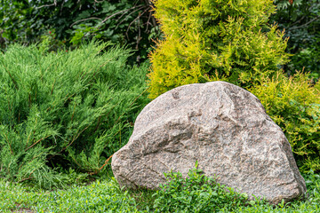 A huge granite stone boulder on a grass lawn against the background with bushes and coniferous trees in a garden design close-up. Beautiful park landscape.