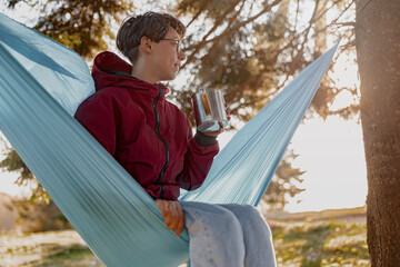 Young woman in glasses sitting in hammock with thermos in forest and enjoying morning.