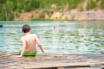 a little boy sits on a wooden bridge after swimming in the lake. wet child sits on the shore of the lake.