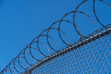 Fence with razor wire with blue sky in the background is shown. 
Razor wire is made up of high tensile core wire and a punched steel tape with sharp barbs at close intervals uniformly.
 - obrazy, fototapety, plakaty