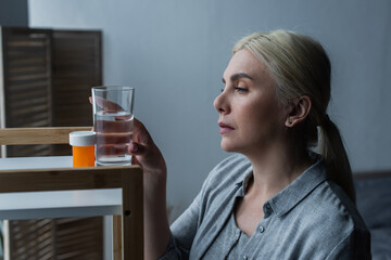 blonde woman with menopause looking at medication and reaching glass of water on table.