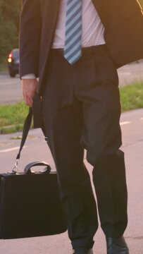 SLOW MOTION, SUN FLARE, LOW ANGLE: Unrecognizable sad man in a suit walking down the street at sunset. Young businessman in despair slowly walking along the sunlit road with his briefcase in hand.