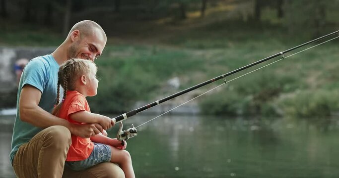Father and little daughter sit on river bank fishing with rod together in park at sunset. Little blonde girl sits on father lap and enjoys fishing