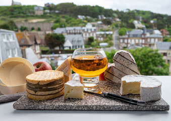 Cow cheeses of Normandy - camembert, livarot, neufchatel, pont l'eveque and glass of apple cider drink with houses of Etretat village on background, Normandy, France