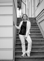 Fashion and beauty concept. Black and white portrait сonfident сaucasian woman in a white suit standing on the stairs, looking into the camera. Background of blue city buildings.
