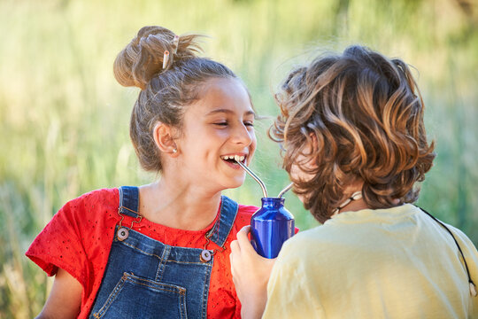 Smiling Girl With Bun And Boy With Curly Hair Drinking Fresh Beverage From Metal Bottle Through Straws And Looking At Each Other During Picnic On Meadow