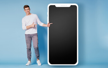 Advertisement. Mobile phone internet concept. Colourful studio portrait of young handsome man pointing big smartphone on the blue background.