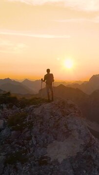 AERIAL, SILHOUETTE, LENS FLARE: Stunning sunset illuminates the Alps and hiker standing on edge of a cliff and observing the picturesque mountain landscape. Man watching the sunrise in the mountains.