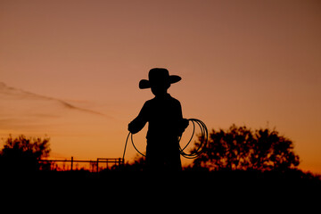 Western industry concept with kid cowboy practicing roping for rodeo at sunset in silhouette on Texas ranch. - obrazy, fototapety, plakaty