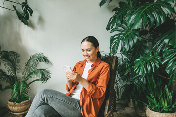 A young smiling woman is sitting with a phone in her hand in a green garden with plants. The concept of remote work or training.