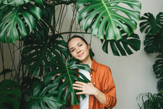 A beautiful young smiling gardener woman stands in her flower shop among green plants. The concept of a green office, plant care, gardening, a modern green recreation area.