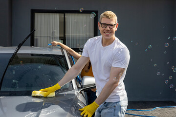 Young Caucasian man is smiling while using sponge and soap water to wash his car in the garage at...
