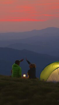 SLOW MOTION: Happy woman and boyfriend high five while sitting by the lantern in the tranquil mountains and enjoy the sights of the beautiful landscape on a cold summer evening. Couple camping in Alps