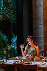 Woman on dinner in luxury modern restaurant. Attractive girl drinking water from glass, looking at window in hotel restaurant. Elegant lady on travel vacation
